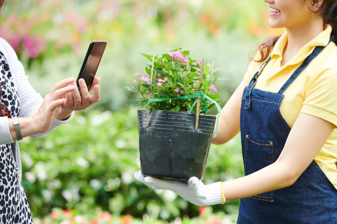 Gardening center worker showing blooming plant to customer who is photographing it on smartphone